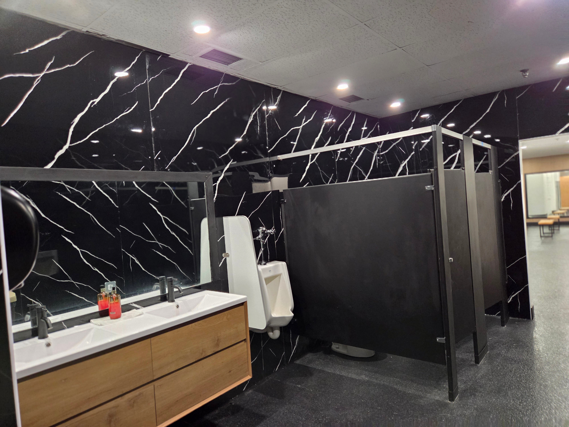 Salle de bain des hommes au Club Fitness à LaSalle, Montréal — double lavabo avec meuble en bois, murs en marbre noir et séparateurs de cabines foncés